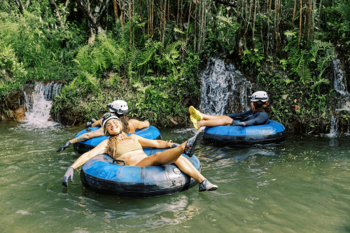 Three people relax on inner tubes in a lush, green river, surrounded by waterfalls and tropical foliage.