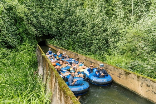 A group of people in blue inner tubes floating down a narrow, green-lined waterway surrounded by lush vegetation.