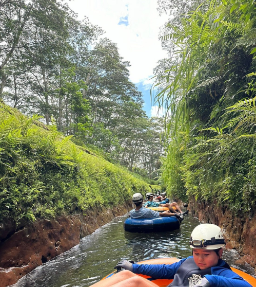 A group of people tubing down a narrow, lush green river surrounded by trees under a bright blue sky.