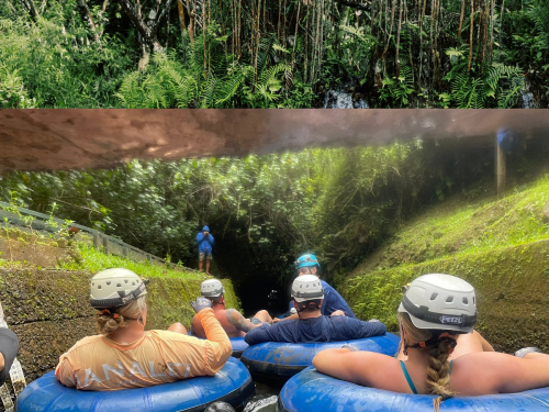 A group of people in helmets float on inner tubes in a lush, green tunnel surrounded by tropical vegetation.