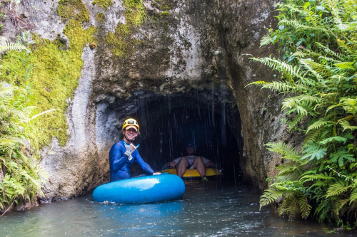 Two people in wetsuits sit in kayaks inside a moss-covered cave, surrounded by ferns and dripping water.