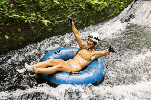 A woman in a bikini joyfully rides a blue inner tube down a flowing stream, surrounded by lush greenery.
