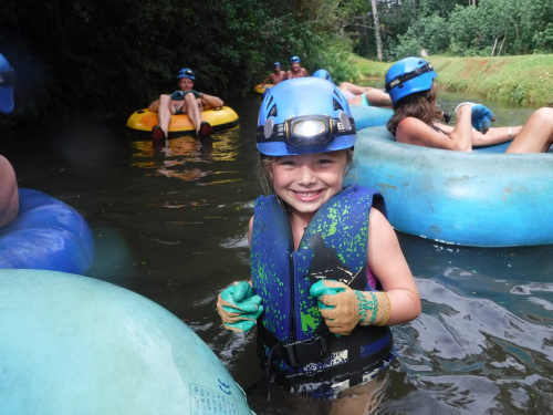 A smiling girl in a life jacket and helmet stands in a river, surrounded by people on inflatable tubes.