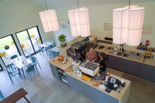A cozy café interior with two baristas at the counter, bright lighting, and seating areas visible in the background.