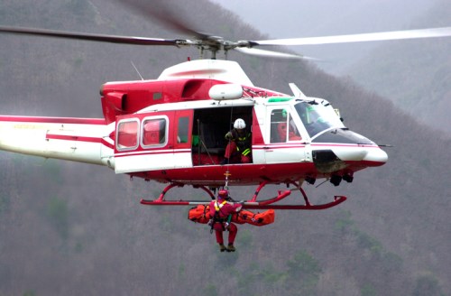 A red and white helicopter hovers above, with a rescuer being lowered on a stretcher to the ground below.