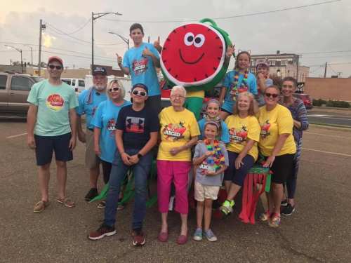 A group of people in colorful shirts poses joyfully around a large, smiling watermelon decoration at a parade.