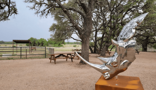 A shiny rhinoceros sculpture stands in a park with trees and picnic tables in the background.