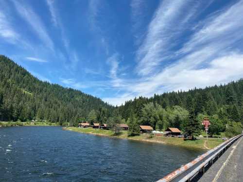 A serene river scene with lush green hills, blue sky, and cabins along the water's edge.