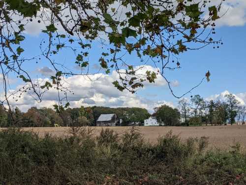 A rural landscape featuring a barn and farmhouse surrounded by fields and trees under a partly cloudy sky.