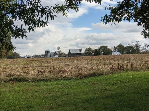 A rural landscape featuring a harvested field, barns, and a silo under a partly cloudy sky.