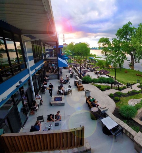 A lively outdoor dining area by a lake, with people enjoying food and drinks as the sun sets in the background.