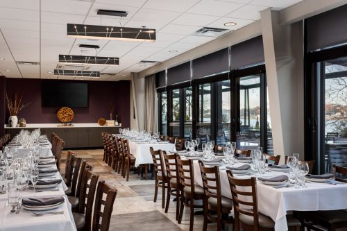 Elegant restaurant interior with long tables set for dining, large windows, and modern lighting fixtures.