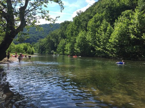 A serene river scene with people kayaking and swimming among lush green trees and mountains under a clear blue sky.