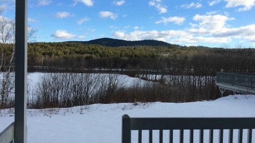 A snowy landscape with a winding river, surrounded by trees and mountains under a partly cloudy sky.