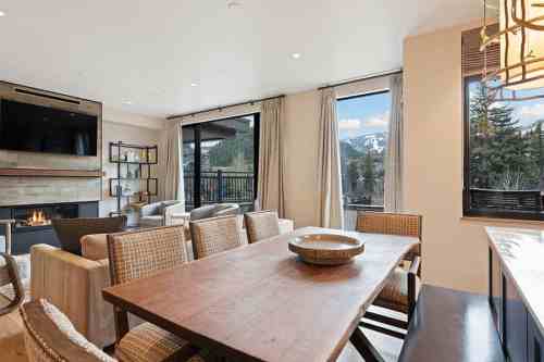 Modern living space with a dining table, cozy seating, large windows, and a view of mountains. Warm, inviting atmosphere.