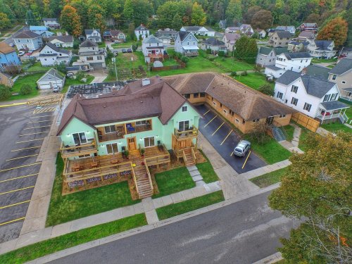 Aerial view of a residential area featuring houses, a green building, and parked cars on a tree-lined street.