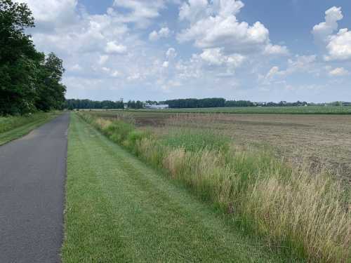 A paved path alongside a grassy area leads to an open field under a partly cloudy sky.