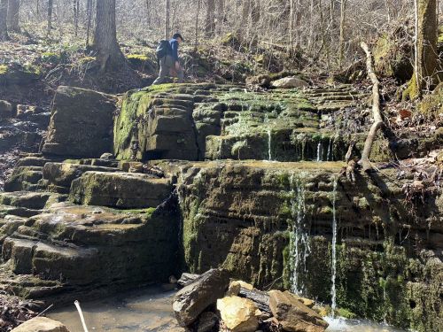 A person explores a moss-covered rock formation with a small waterfall in a wooded area.