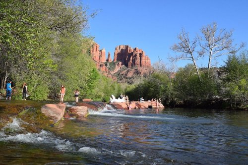 A serene river scene with people enjoying nature, surrounded by lush greenery and red rock formations under a clear blue sky.