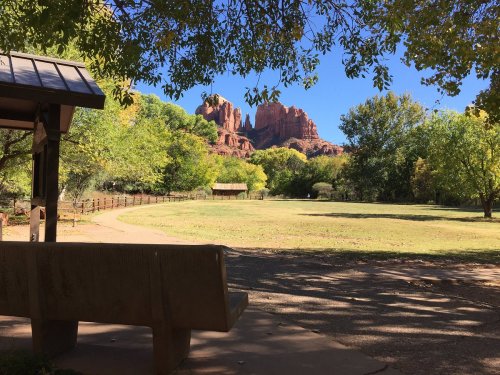 A serene park scene with a bench, green trees, and red rock formations in the background under a clear blue sky.