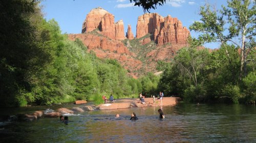 A serene river scene with people swimming, surrounded by lush greenery and towering red rock formations in the background.