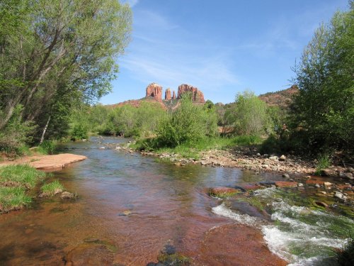 A serene river flows through lush greenery, with red rock formations rising in the background under a clear blue sky.