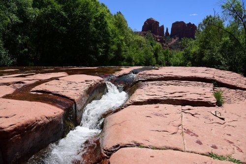 A serene river flows over smooth red rocks, surrounded by lush greenery and distant rock formations under a clear blue sky.