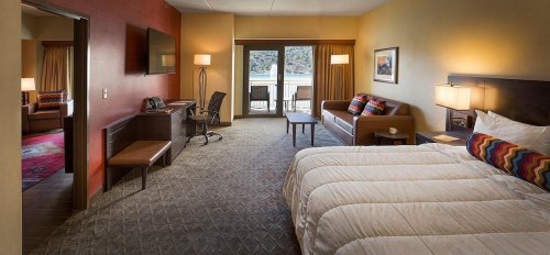 A spacious hotel room featuring a bed, seating area, desk, and balcony with a view. Warm colors and modern decor.