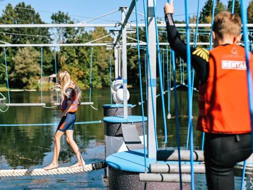 A girl walks on a rope bridge over water while a boy in a life jacket observes from the side.