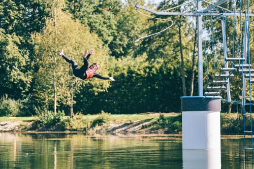A person in a red life jacket jumps into a lake from a platform, surrounded by trees and greenery.