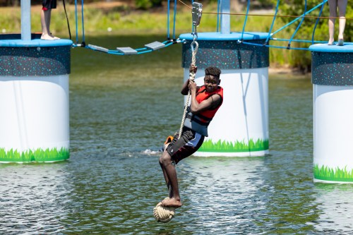 A person in a life jacket swings on a rope over water, preparing to land on a floating platform.