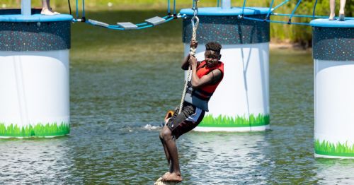 A person in a life jacket swings on a rope over water, approaching a colorful floating structure.