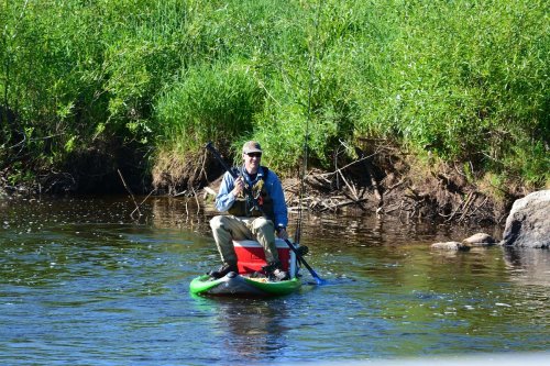 A person fishing from a kayak on a calm river, surrounded by lush green grass and trees.