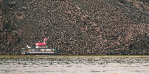 A houseboat on a river, featuring a large pink flamingo float on its roof, with rocky terrain in the background.