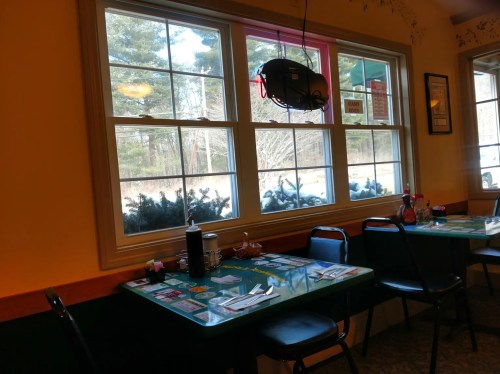 A cozy restaurant interior with a green table, chairs, and large windows showing a snowy landscape outside.
