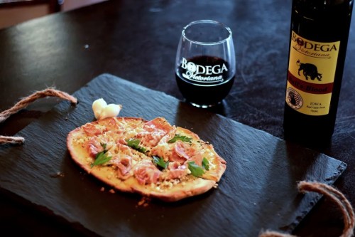 A flatbread topped with ham and herbs, served on a slate plate with a glass of red wine and a bottle in the background.
