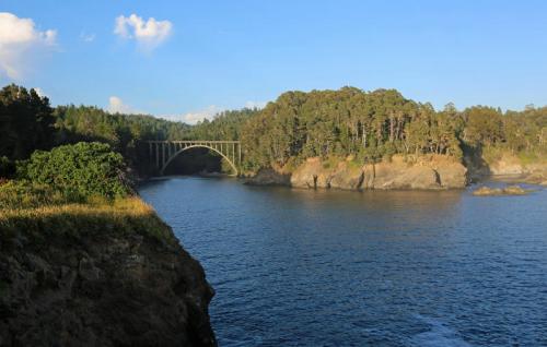 A scenic view of a bridge over a calm river, surrounded by lush greenery and rocky cliffs under a clear blue sky.
