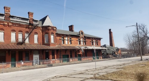 Abandoned red brick building with a collapsed roof, surrounded by a fence and a clear blue sky.