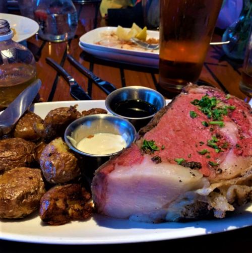A plate featuring a large slice of prime rib, roasted potatoes, and dipping sauces, set on a wooden table.