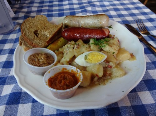 A plate of sausages, potato salad, boiled egg, mustard, and bread on a blue and white checkered tablecloth.