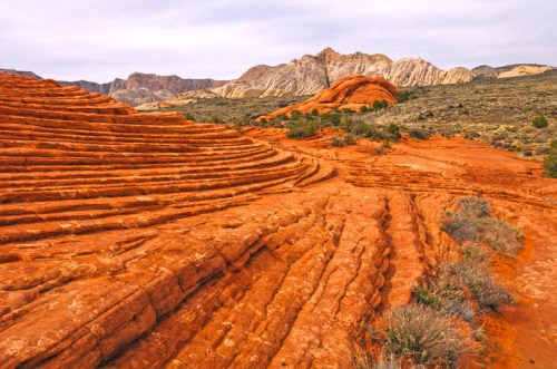 Layered red rock formations with green shrubs, set against a backdrop of mountains under a cloudy sky.