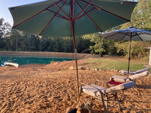 Two lounge chairs under large umbrellas by a serene pond, surrounded by trees and sandy ground.