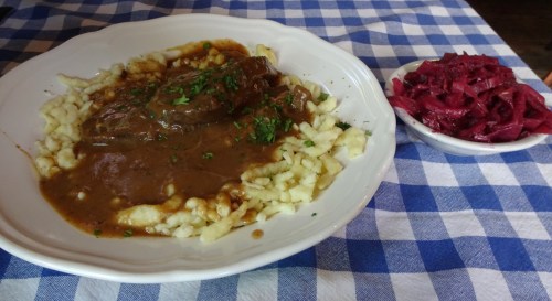 A plate of meat with gravy over dumplings, garnished with parsley, alongside a bowl of red cabbage on a checkered tablecloth.