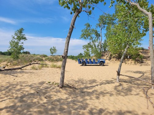 A blue vehicle parked on sandy terrain surrounded by sparse trees and a clear blue sky.