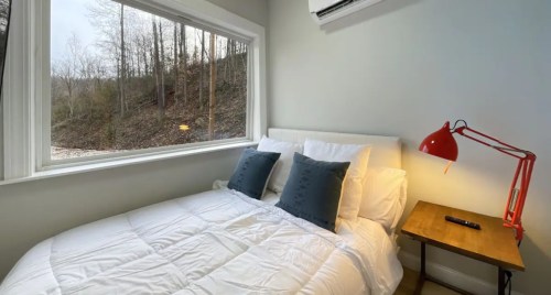Cozy bedroom with a large window, a bed with white linens, blue pillows, and a red lamp on a wooden nightstand.