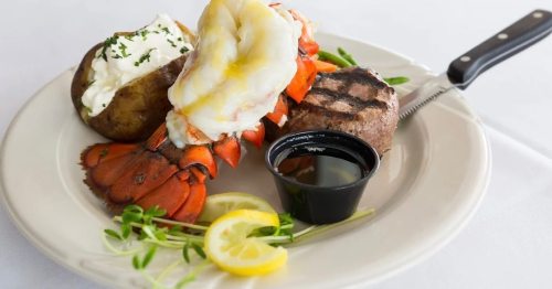 A plate featuring grilled lobster tail, steak, a baked potato with sour cream, and lemon slices.