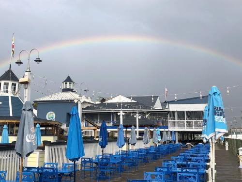 A waterfront scene with blue chairs and umbrellas, under a rainbow and cloudy sky. Buildings in the background.