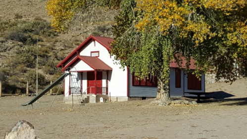 A small white house with a red roof, surrounded by trees and a slide, set in a dry, open landscape.
