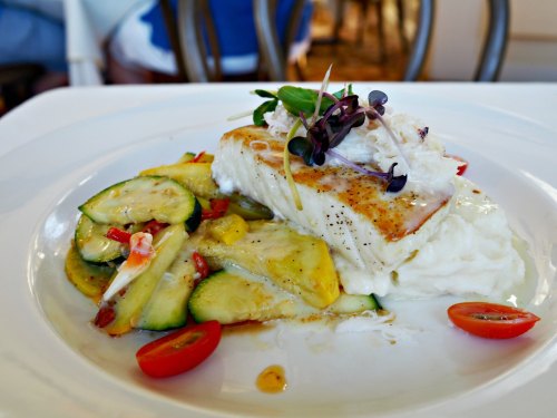 A plated dish featuring grilled fish on a bed of mashed potatoes, accompanied by sautéed vegetables and cherry tomatoes.