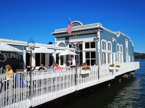 A waterfront restaurant with outdoor seating, people dining, and an American flag flying on a clear blue day.